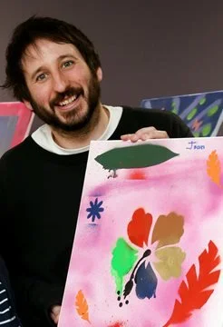 Man with dark hair and beard smiling, holding colorful painted artwork featuring a blimp, flowers, and abstract shapes.