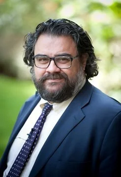 A man with dark glasses, wavy dark hair, and a beard, wearing a navy suit, white shirt, and patterned tie, standing outdoors with greenery in the background.