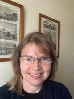 A portrait of a woman with brown hair and glasses. She is wearing a dark top and is stood in front of two framed pictures.