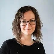 A woman with curly hair and glasses smiling at the camera, wearing a black top, against a light background.