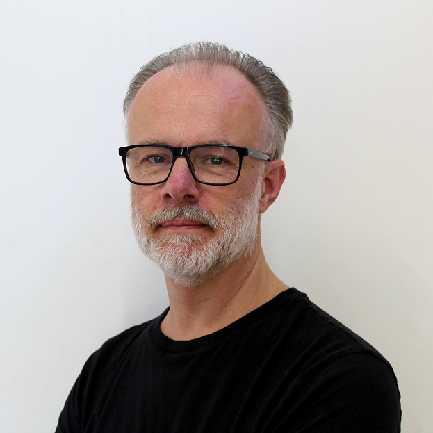 A man with gray hair, a beard, and glasses, wearing a black shirt, standing against a white background.