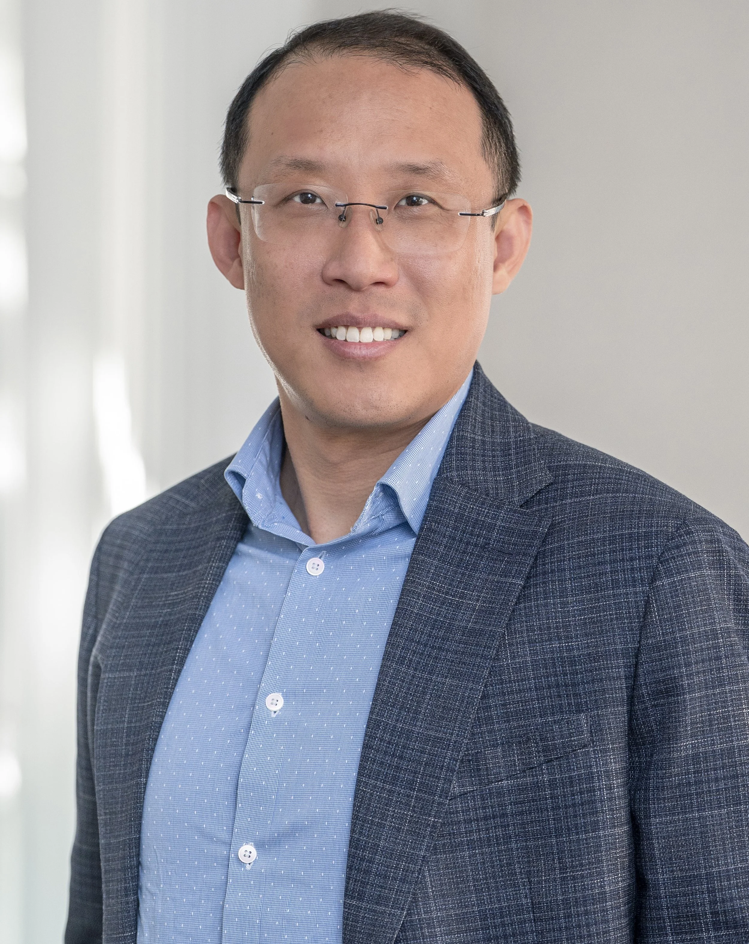 A man wearing glasses, a blue button up shirt and grey blazer. He is smiling for the photo.