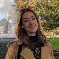 Woman outdoors with trees, wearing a tan coat and red bow headband, smiling.
