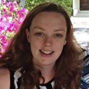 A woman with long brown hair smiling at the camera, indoors with a window and pink flowers in the background.