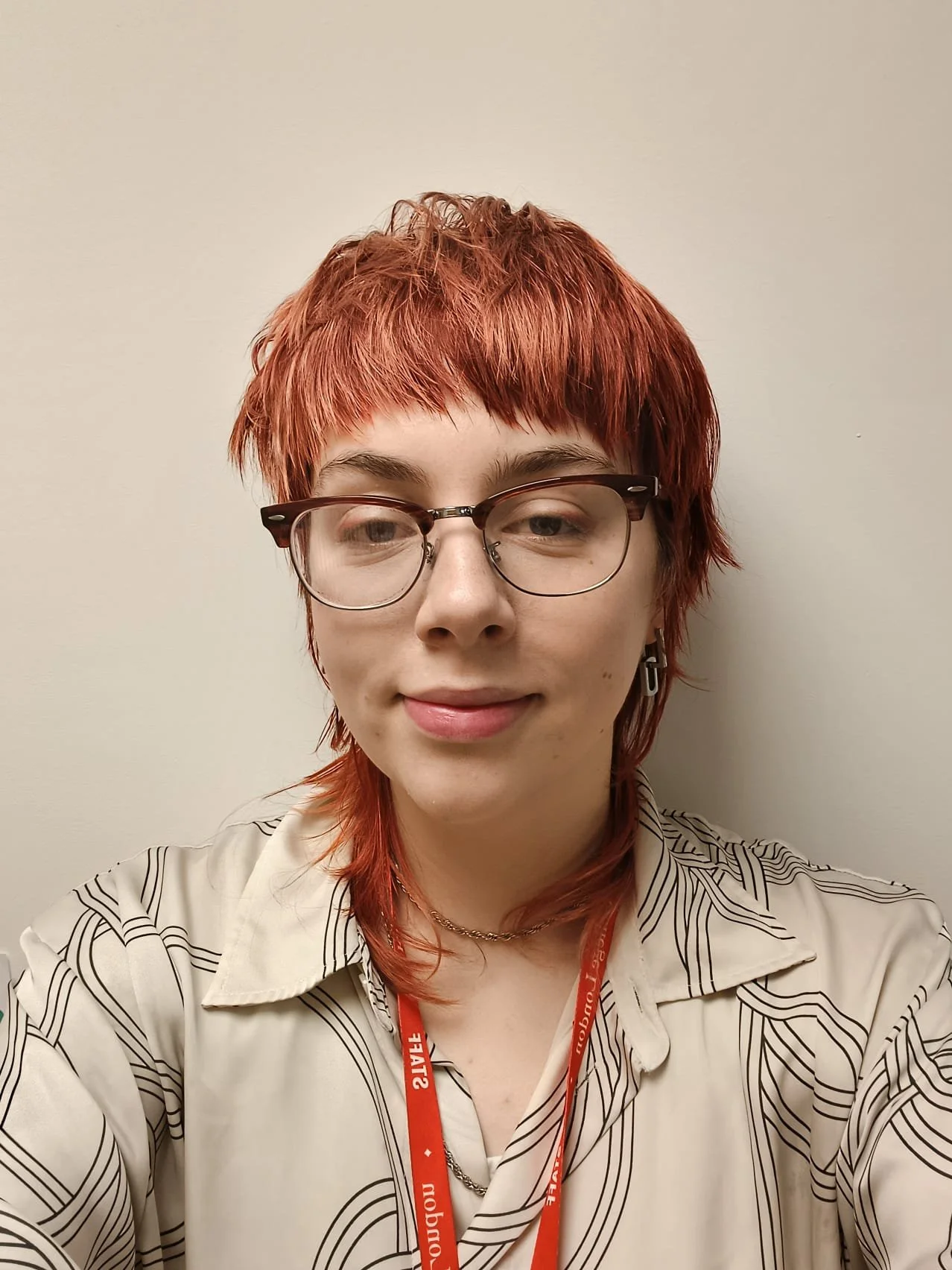A person with red hair standing in front of a plain background smiling. They are wearing glasses and a cream button up shirt