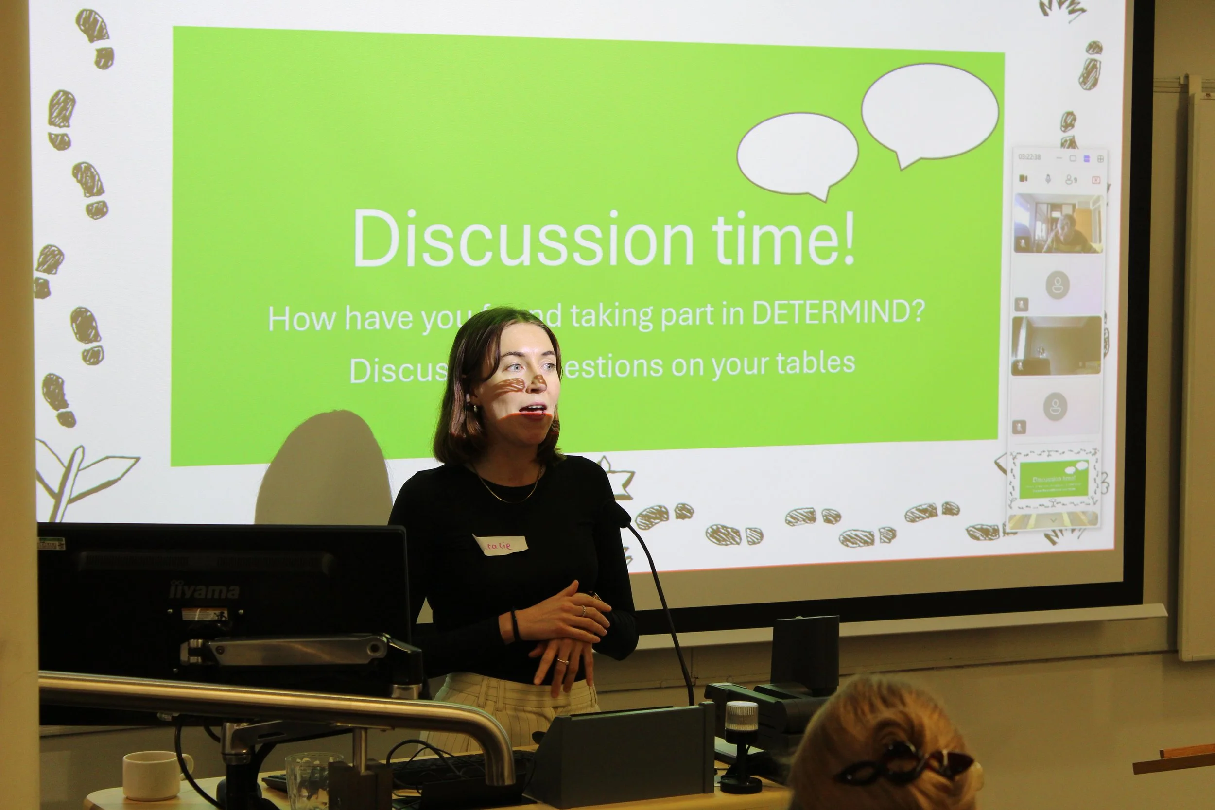 One of the DETERMIND research assistants presenting in front of a projector screen. On the screen, they have a slide named 'Discussion Time', asking people how they found taking part in DETERMIND. The slide is bright with speech bubble illustrations.