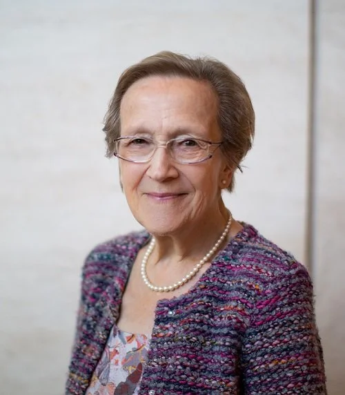 A portrait of a woman with short gray hair and glasses, wearing a pearl necklace and a colorful, textured jacket.