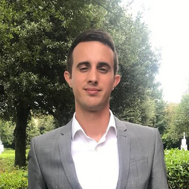 A man in a gray suit and white shirt standing outdoors with trees and greenery in the background.