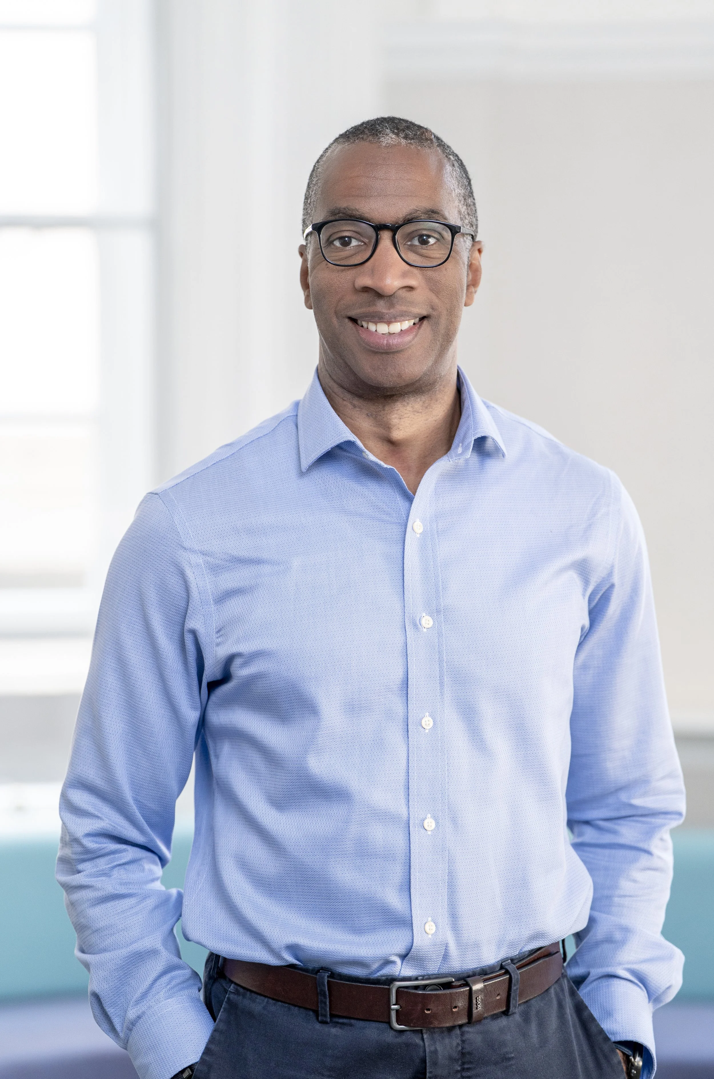 A man wearing glasses and a long-sleeved blue button up shirt. He is smiling for the photo and has his hands in his pockets.