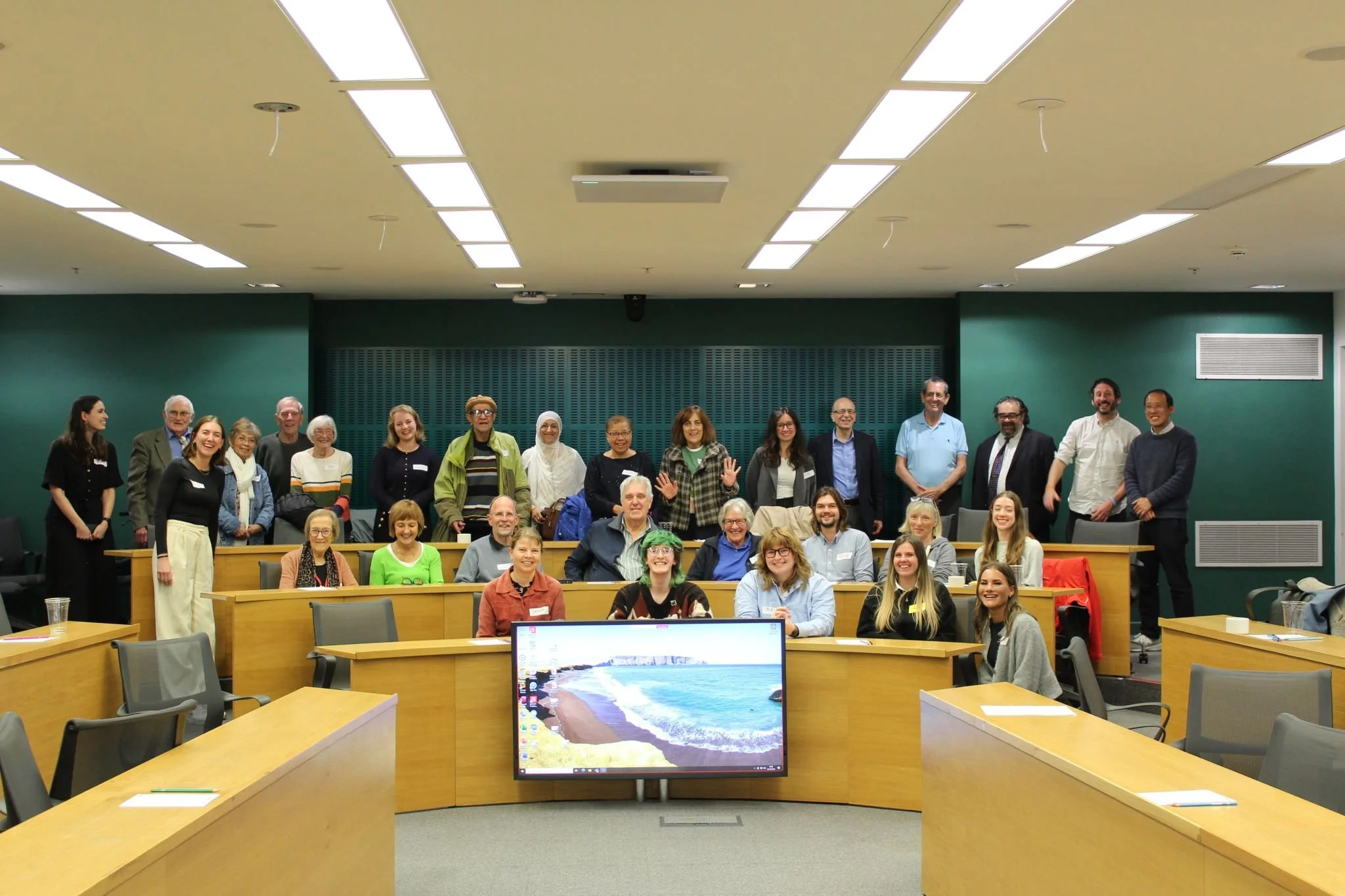 A large group of diverse people gathered in a conference room, posing for a group photo with some standing and others sitting at the tables.