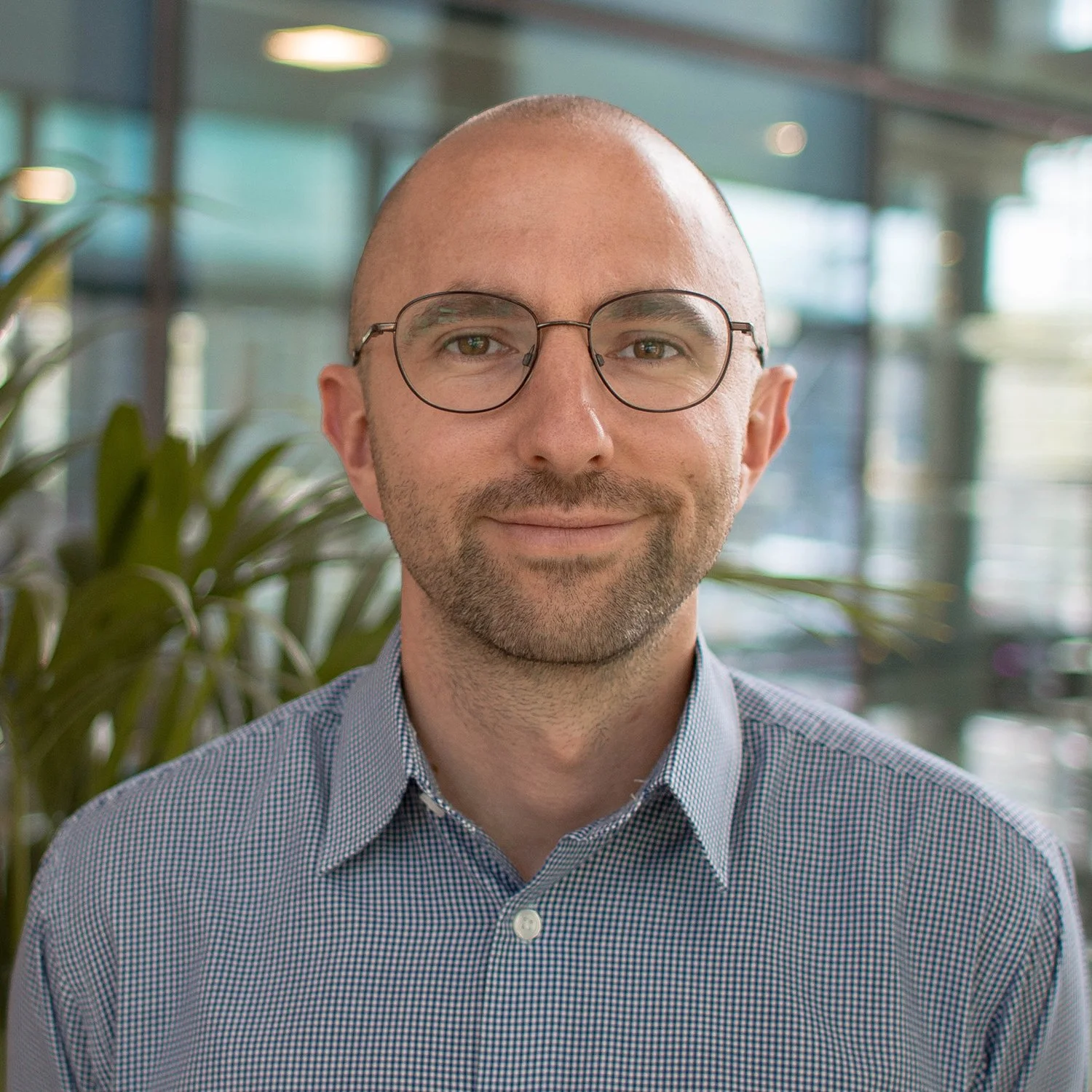 A man with glasses, a beard, and a shaved head smiling in a modern indoor setting with large windows and green plants.