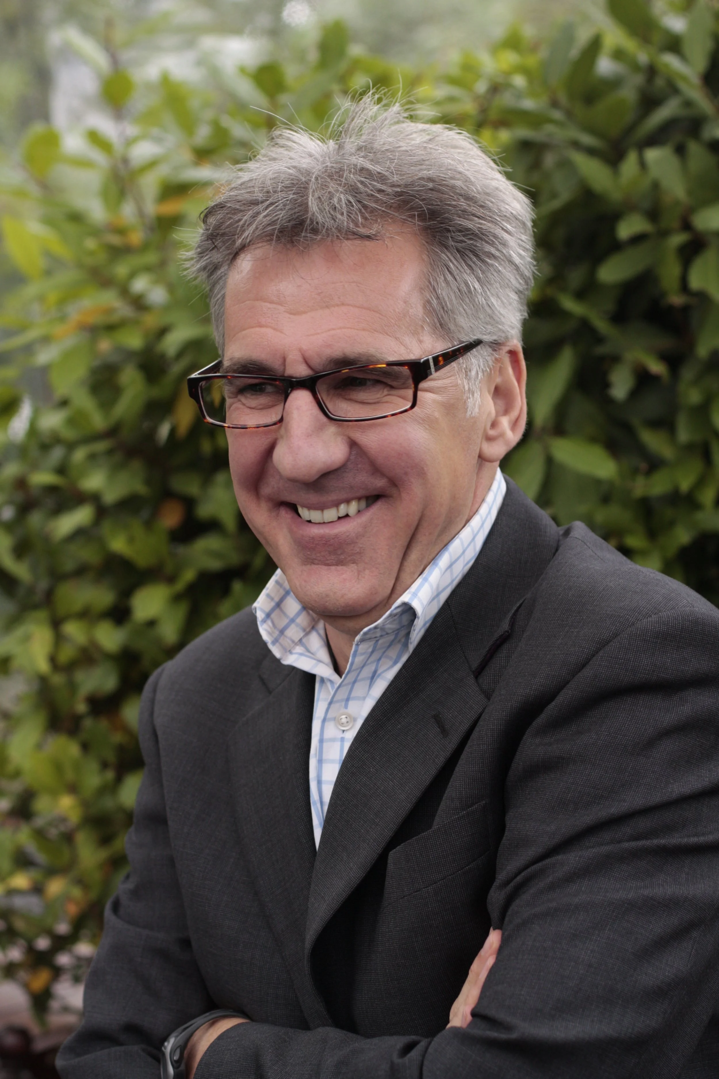 A man in glasses and a gray suit, smiling with his arms crossed, standing outdoors in front of green leafy plants.