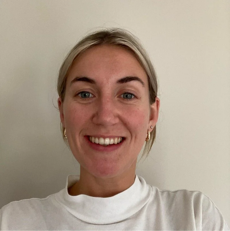A woman with short blonde hair, blue eyes, and a broad smile, wearing a white turtleneck top and gold hoop earrings, standing against a plain light-colored wall.