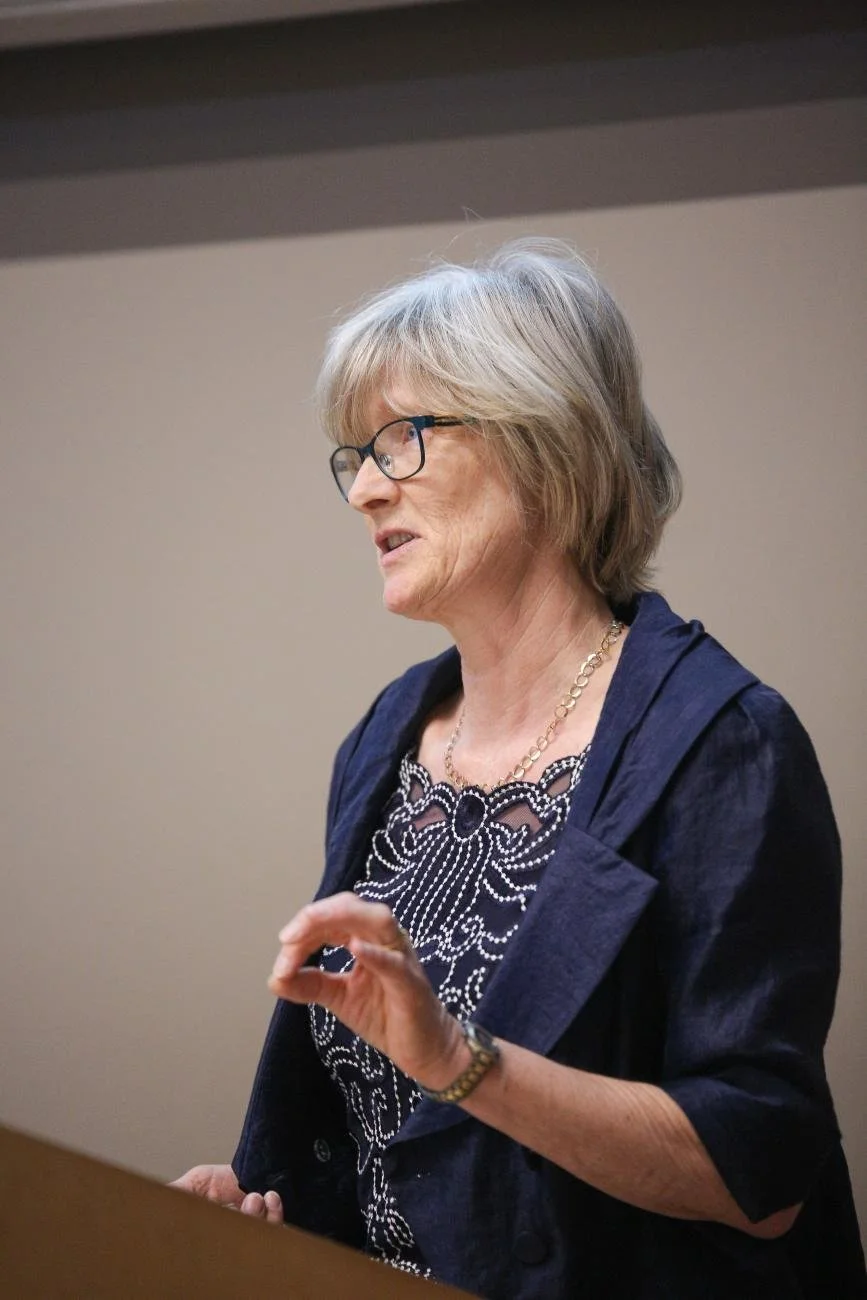 A woman with short gray hair, glasses, and a navy blazer giving a presentation or speech in an indoor setting.