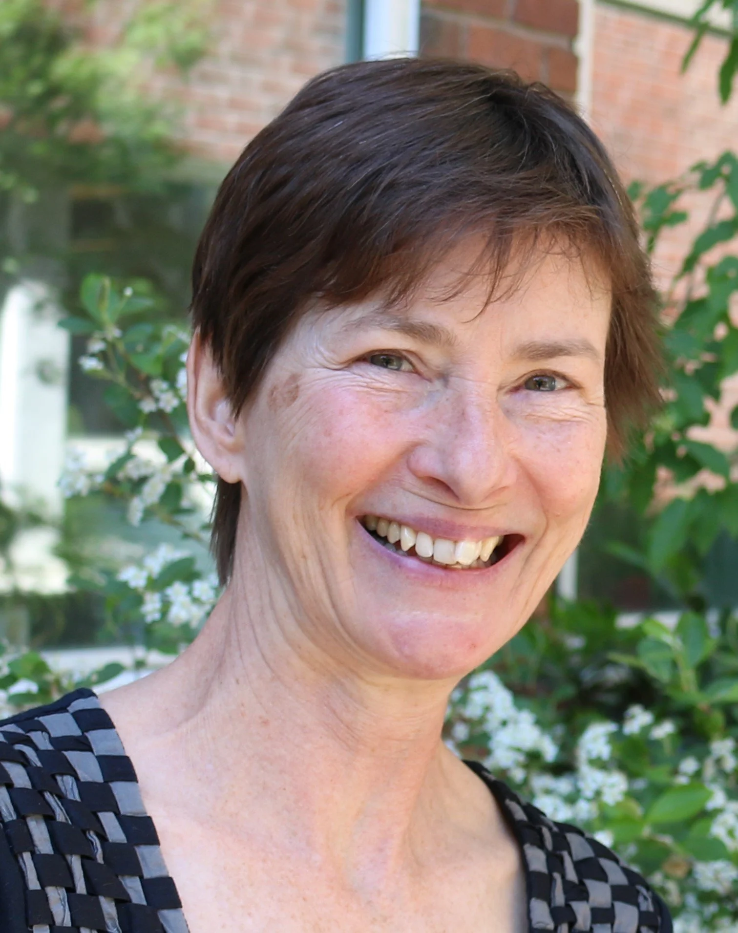 A smiling woman with short brown hair outdoors, with greenery and a brick building in the background.