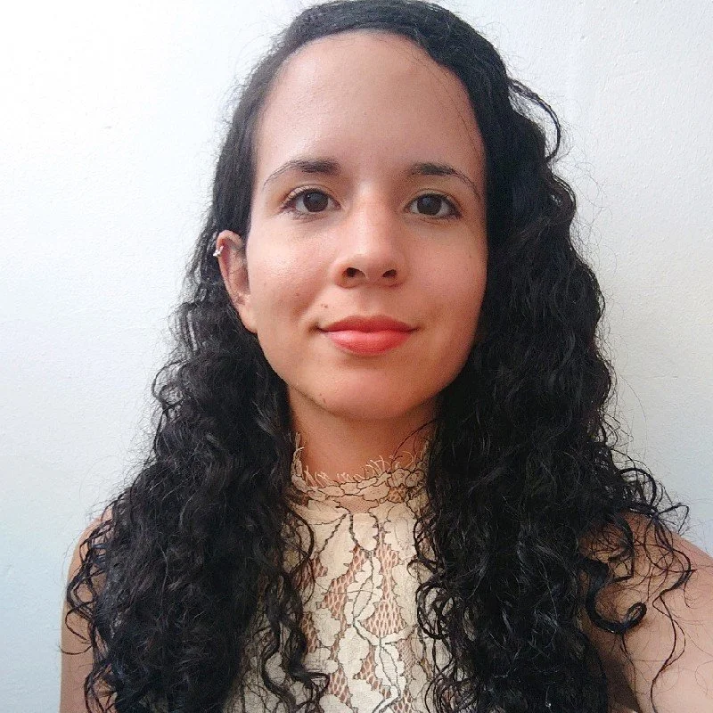 A woman with long, curly dark hair, wearing a beige lace top, smiling slightly, standing against a plain white wall.