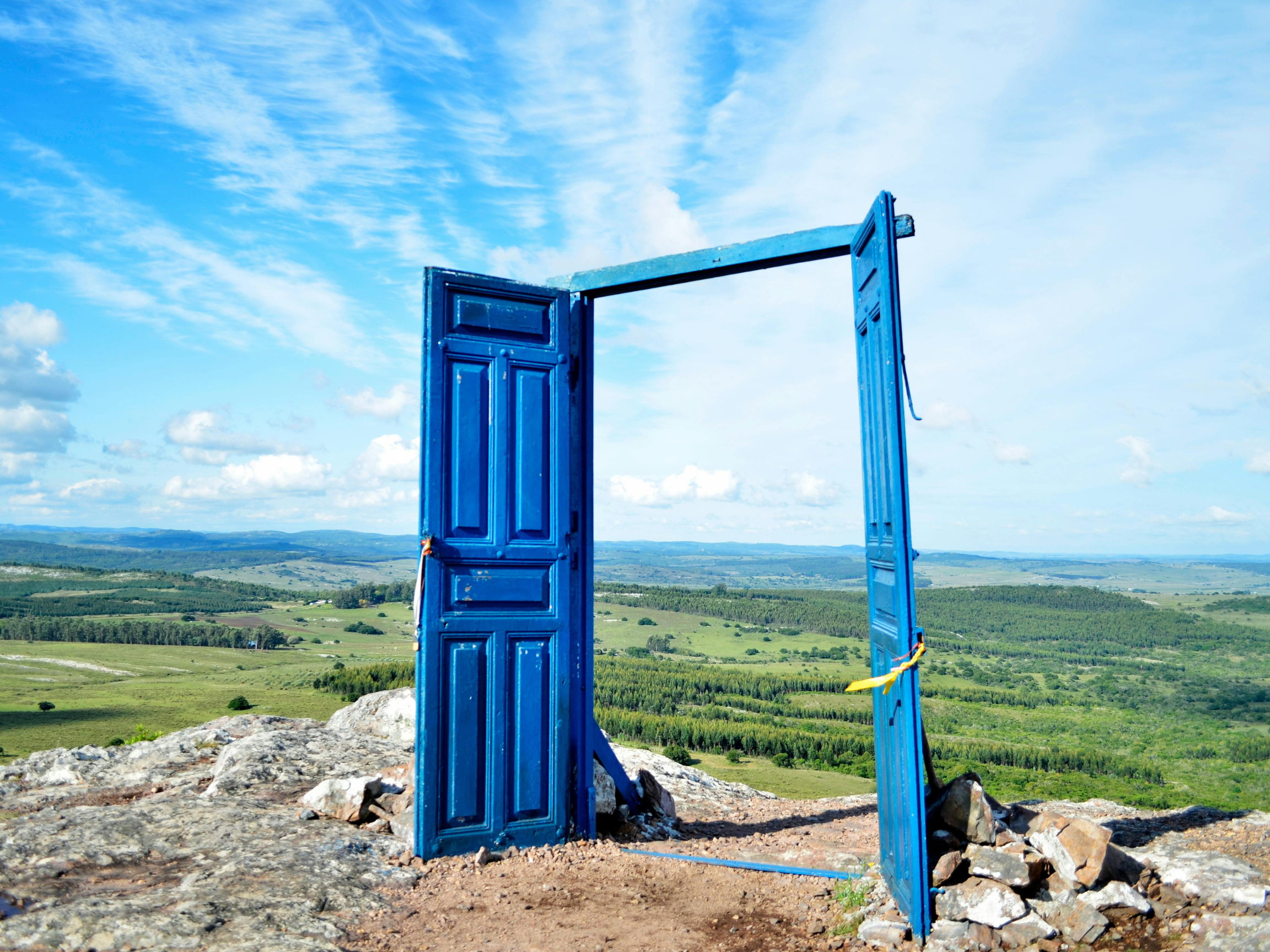 Blue doors standing open on a rocky hilltop overlooking a green valley and blue sky with clouds.