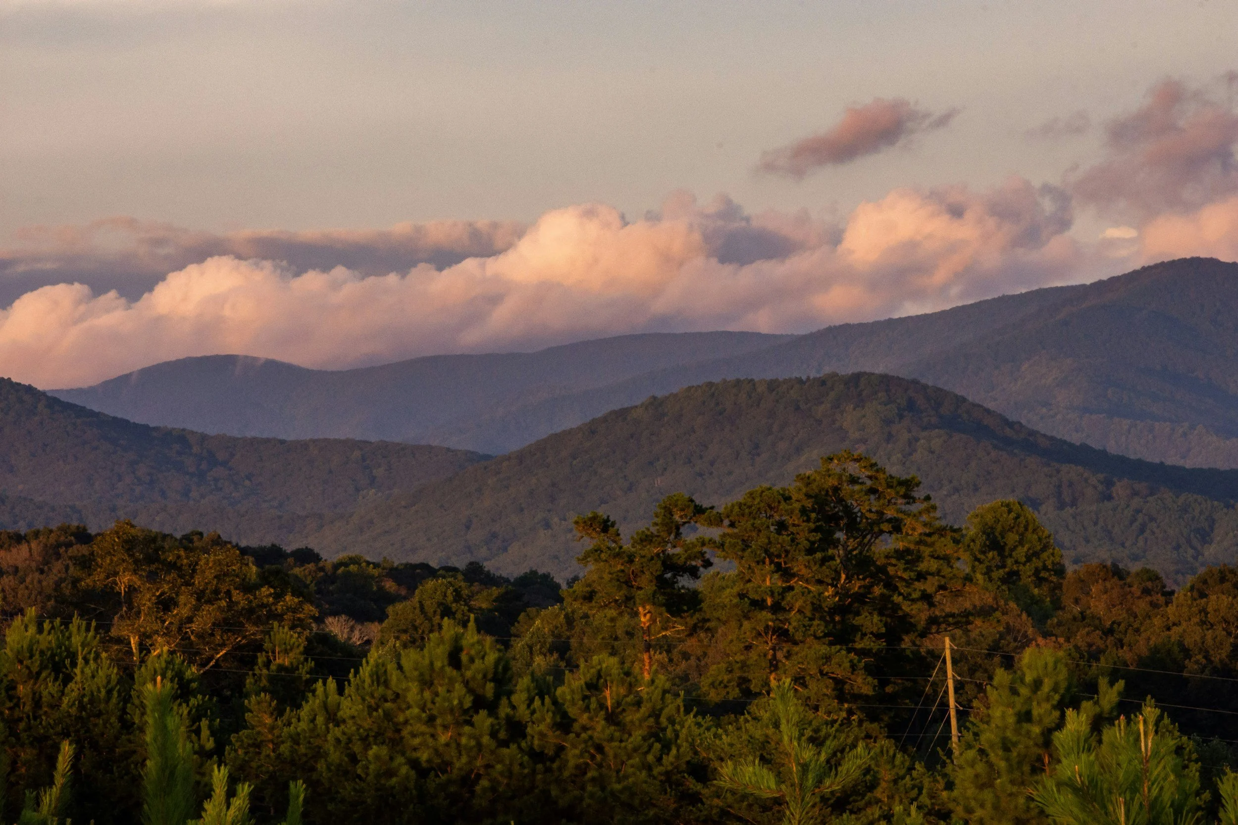 Scenic view of multiple mountain ranges with a forested foreground and clouds in the sky during sunset.
