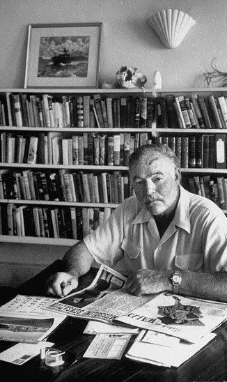 A man sitting at a cluttered desk in a room filled with bookshelves, with a framed picture and decorative items on the wall behind him.