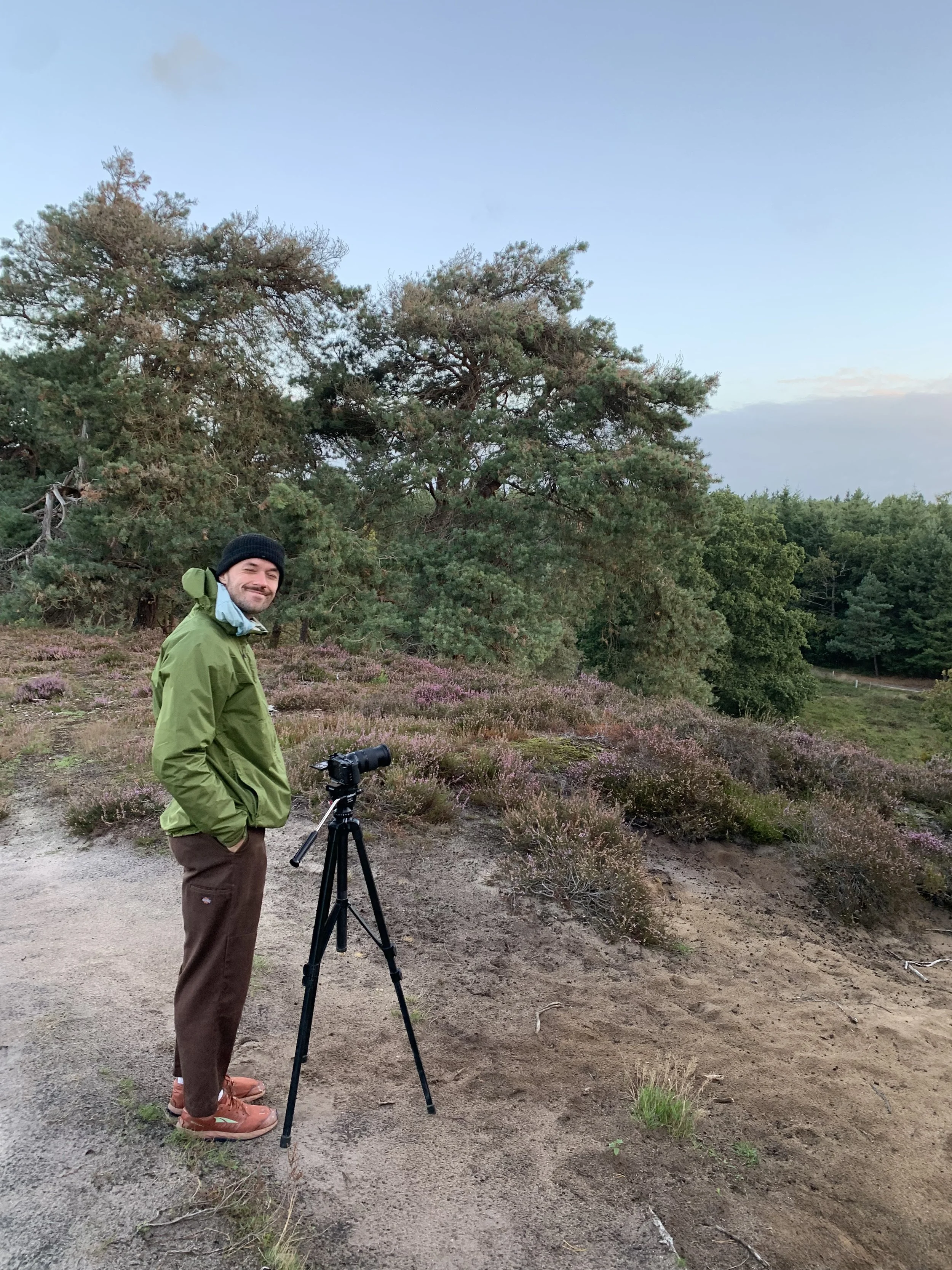 A man stands outdoors on a dirt path with a camera on a tripod, surrounded by trees and purple heather plants.
