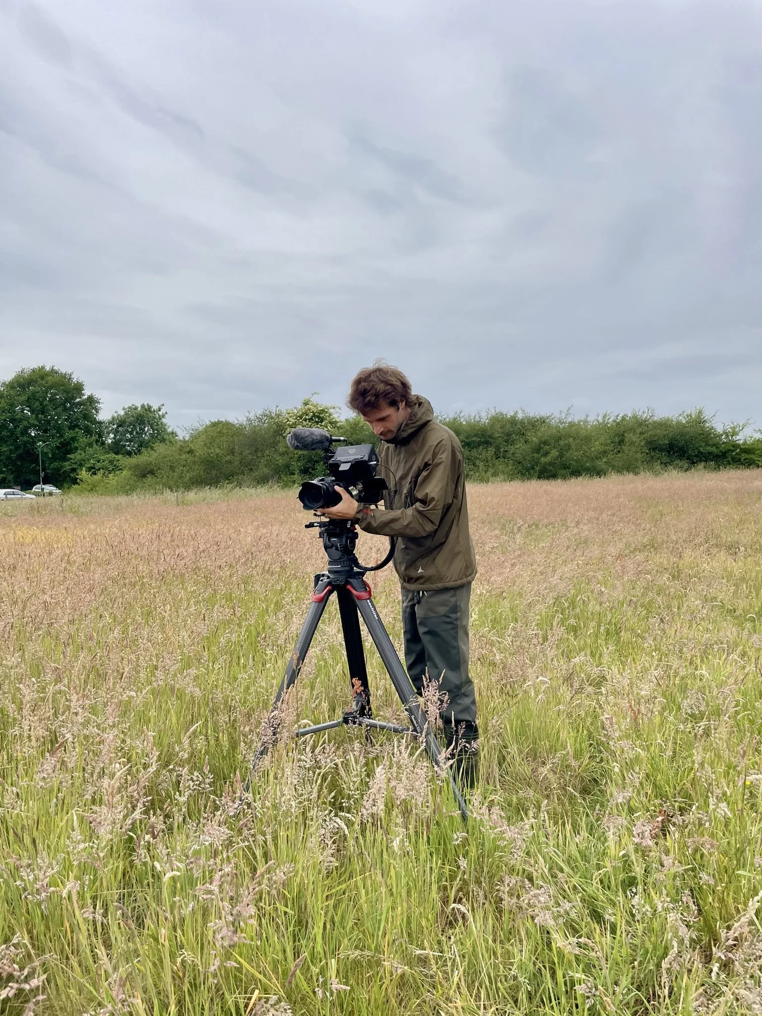 A man operating a professional video camera on a tripod in a grassy field under a cloudy sky.
