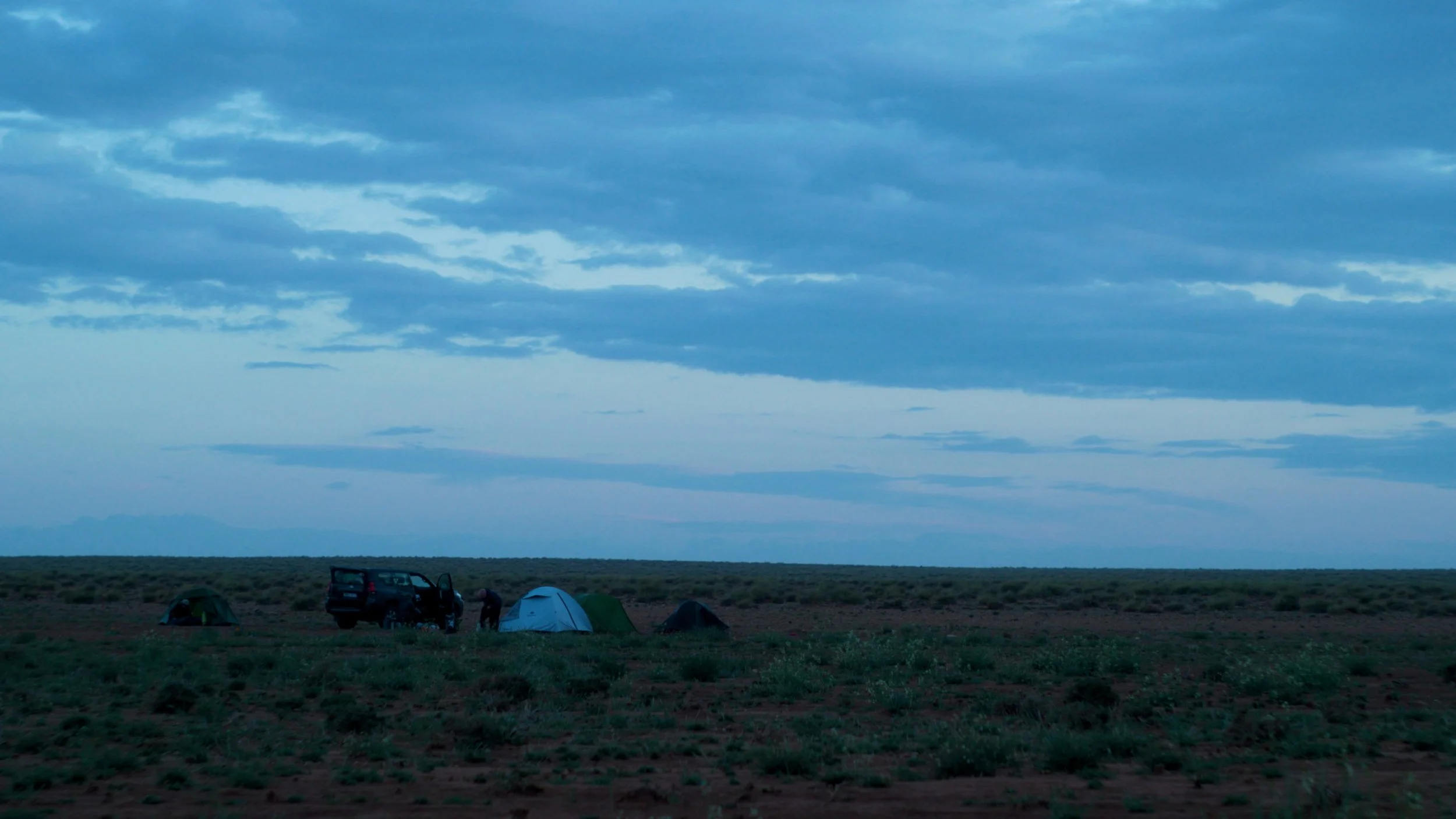 Camping tents and a vehicle set up in a vast open landscape at dusk under a cloudy sky.