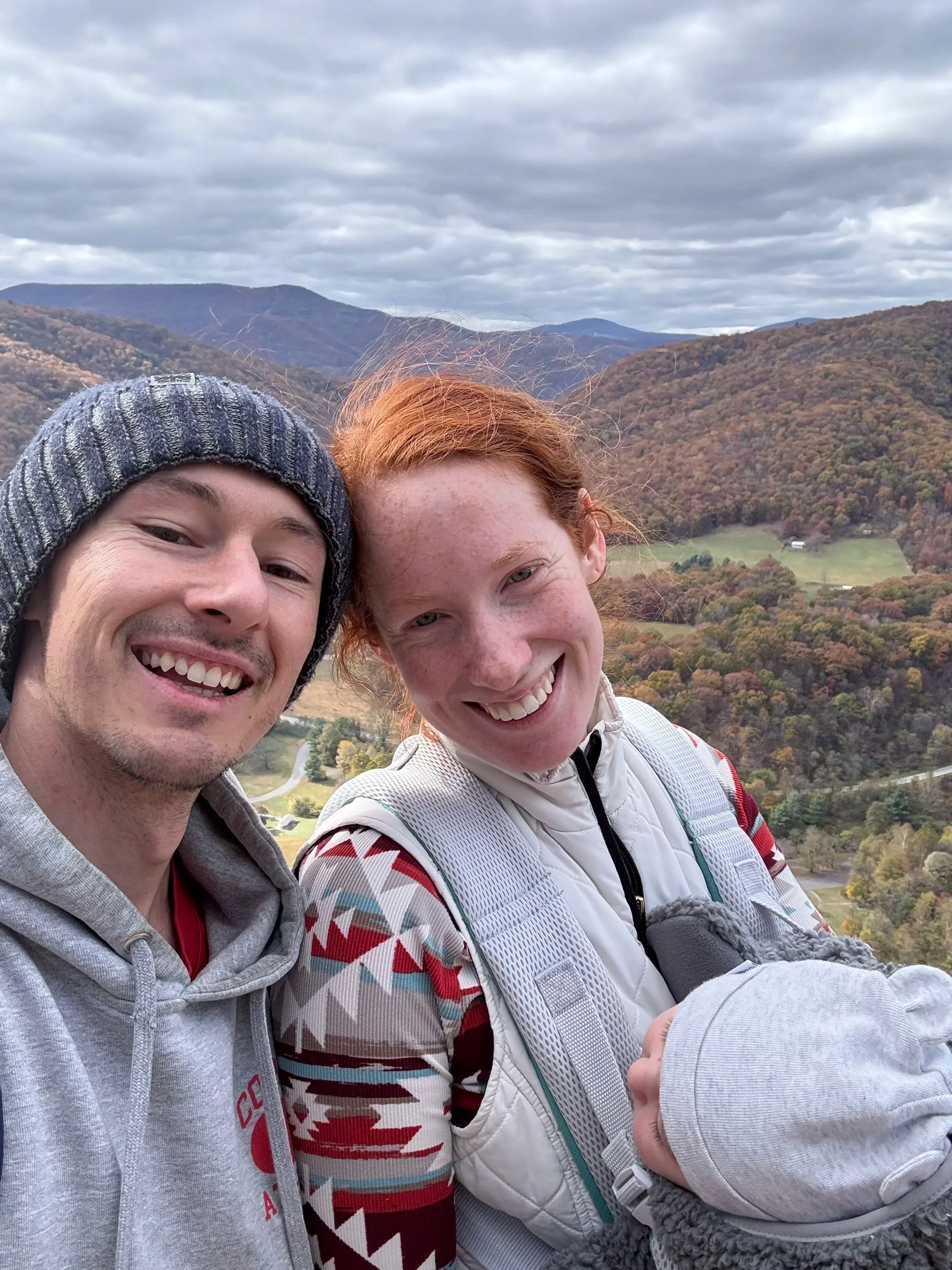 Tyler and Kelly (Eryvalen founders), with their daughter Driscoll, smiling outdoors with a mountainous landscape in the background, showing cloudy sky, rolling hills, and trees with fall foliage.