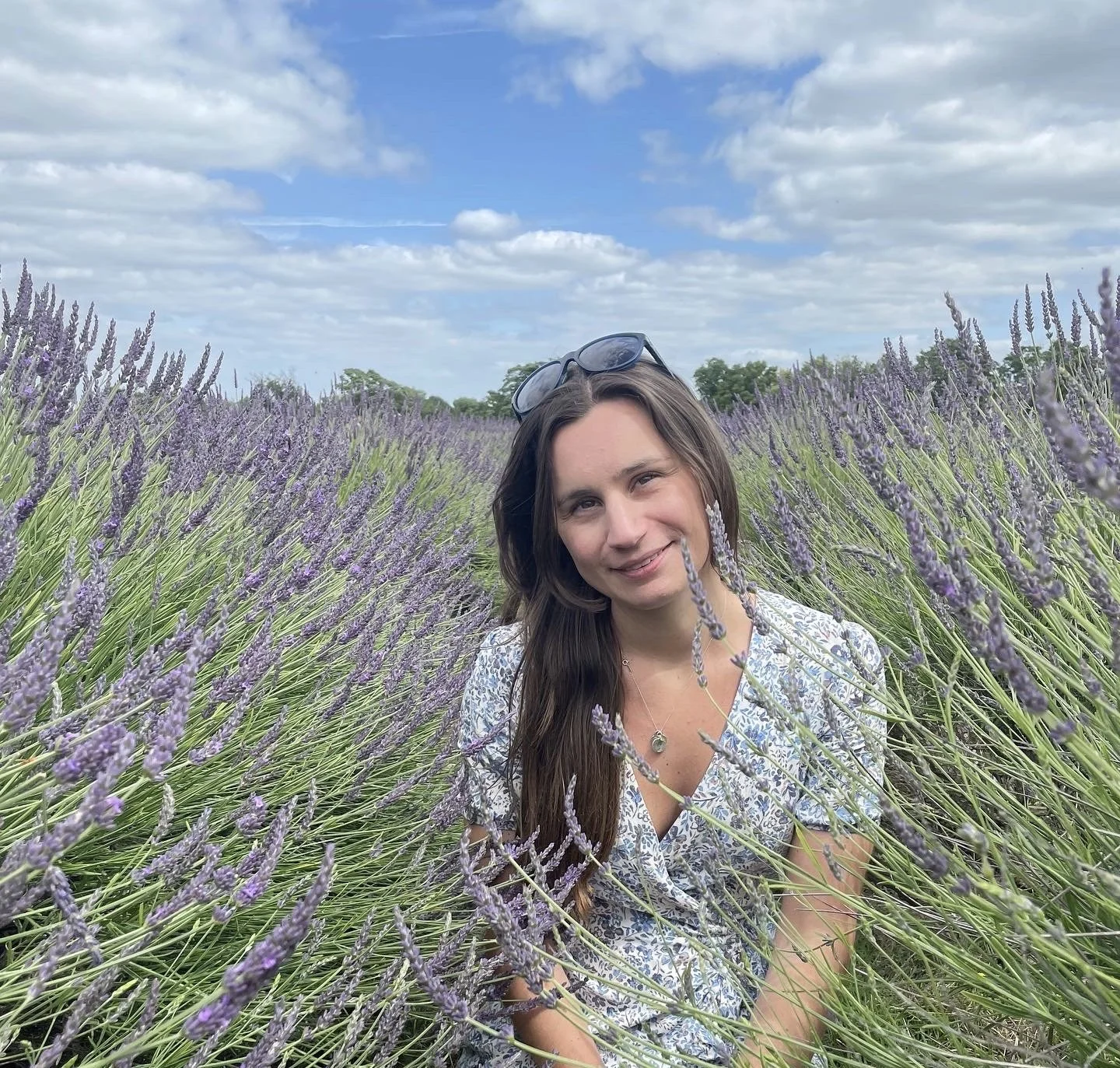 Sophie Redgrave. A woman with long brown hair and sunglasses resting on her head, smiling while sitting among rows of blooming lavender flowers outdoors on a partly cloudy day.