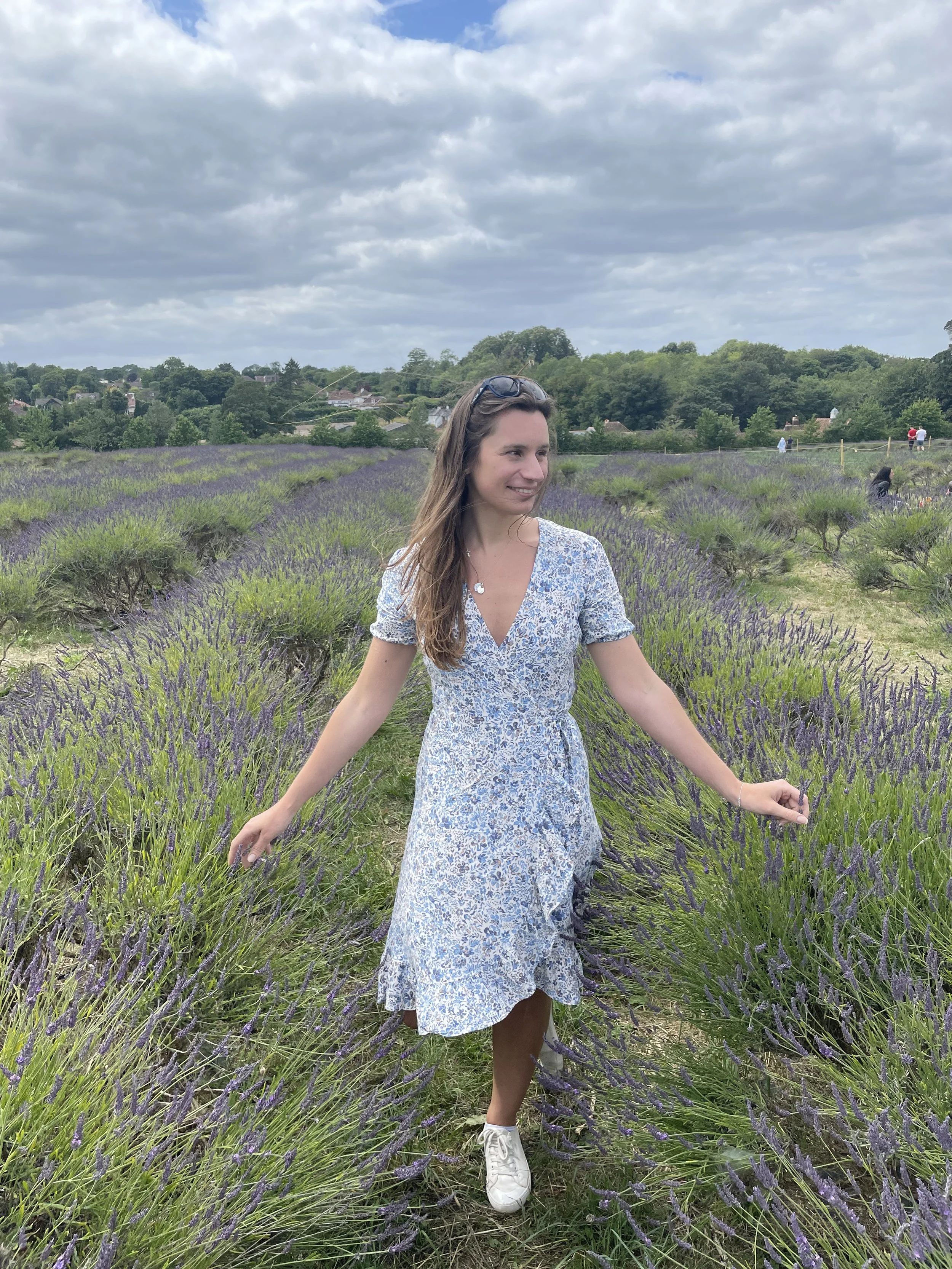 Sophie Redgrave. A young woman wearing a light blue floral dress and white sneakers standing in a lavender field under a cloudy sky, smiling with long hair and sunglasses on her head.