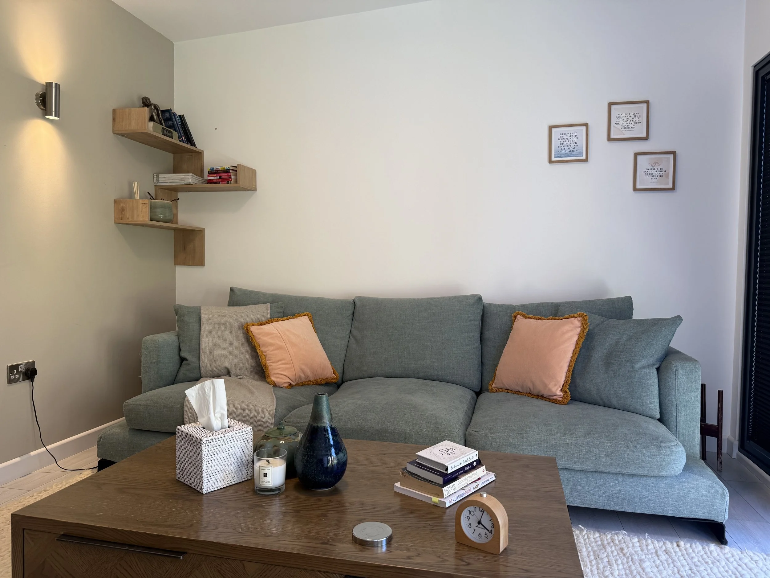 A therapy room that feels like home, a comforting green sofa with peach-colored pillows, a wooden coffee table with decorative vases, a tissue box, candles, a stack of books, and a clock. Soft images and posters on the wall, shelves with books.