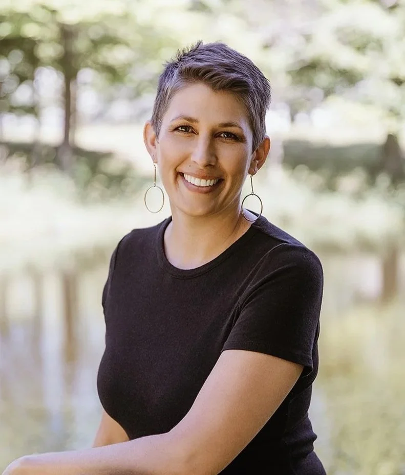 Liza Annette, therapist, with short hair and hoop earrings smiling outdoors with trees in the background.