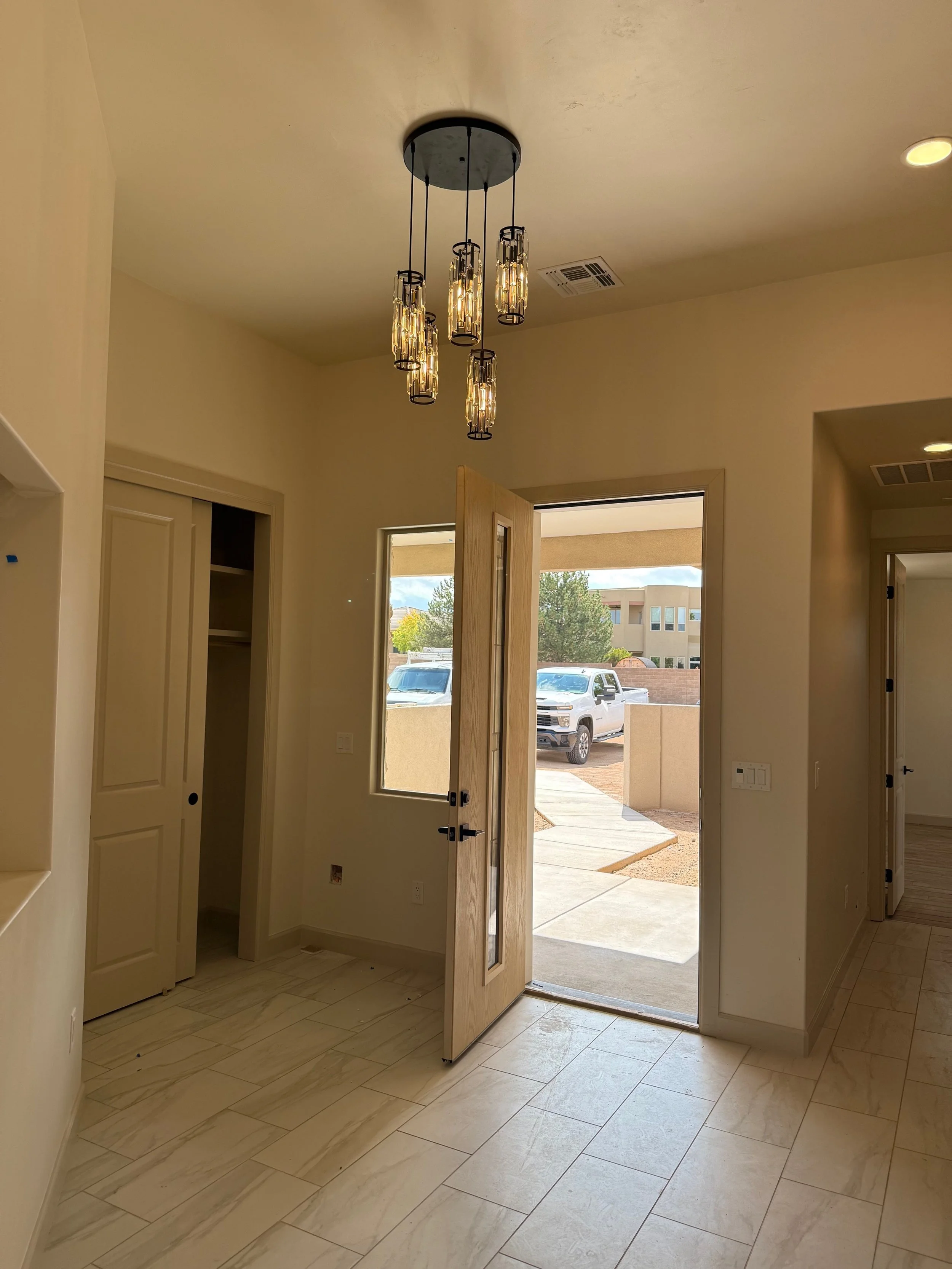 Interior view of a house entryway with a hanging chandelier, open wooden door leading outside, closet with open door, and tiled floor.