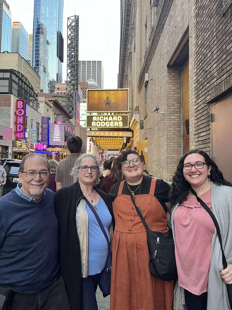 Eden and their family smiling and standing on a busy city street, with bright signs and theater marquee with Richard Rodgers and Hamilton in the background.