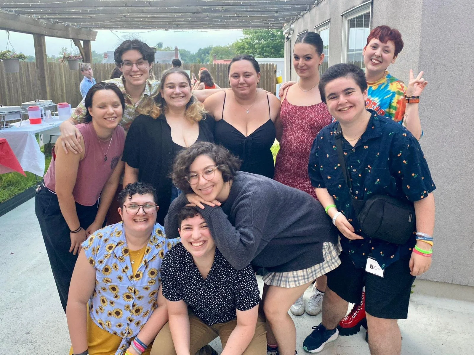 Group of people smiling and posing for a photo at an outdoor gathering, with some in casual clothing and others in party attire, on a patio with a table with food and drinks in the background.