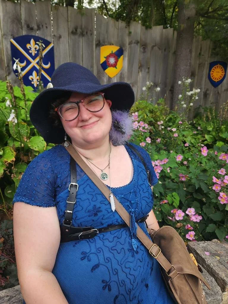 Eden smiling while wearing a blue woven witches hat, glasses, and a blue embroidered dress with a brown strap bag, standing in front of a garden with pink flowers and a wooden fence decorated with heraldic shields.