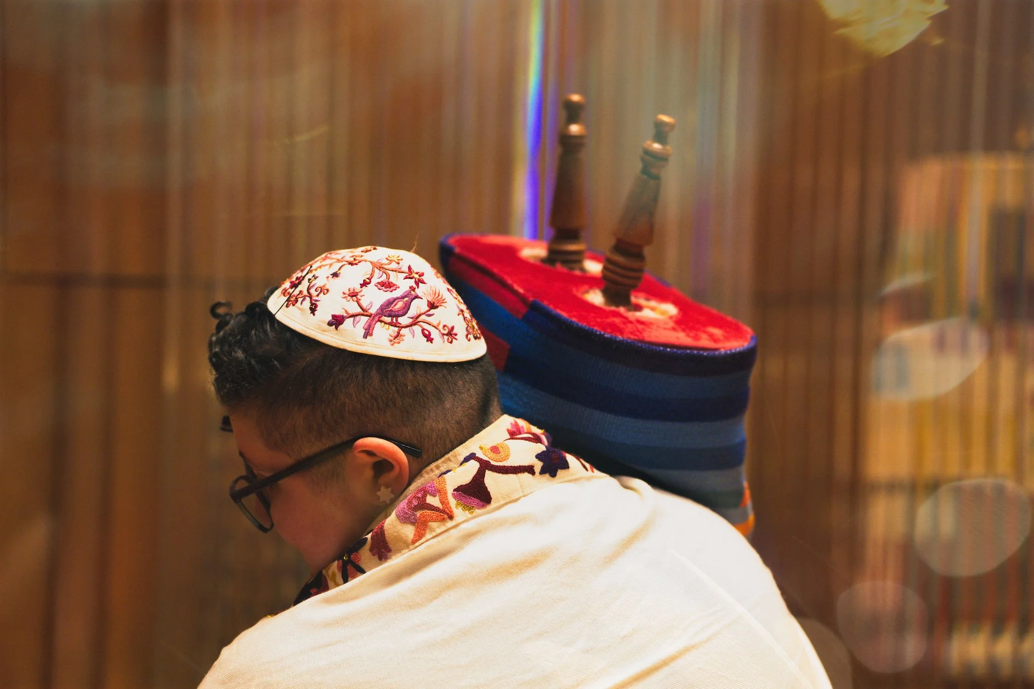 Eden wearing a kippah and sitting with a decorated tallit, returning the Torah in a rainbow mantel to the ark, in front of a wooden background.