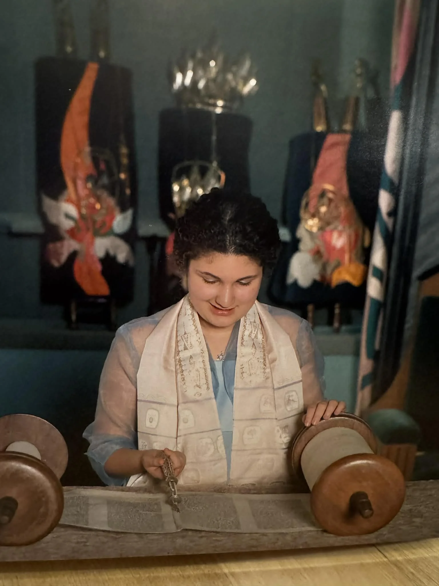 Eden at their bat mitzvah, wearing a cream tallit, prayer shawl, reading a Torah and holding a small yad while smiling. Behind them, colorful fabric covers the ark.