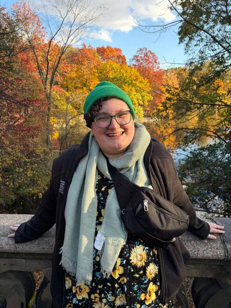 Eden smiling with glasses, green beanie, and light-colored scarf, standing outdoors on a stone railing with colorful autumn trees and blue sky in the background.