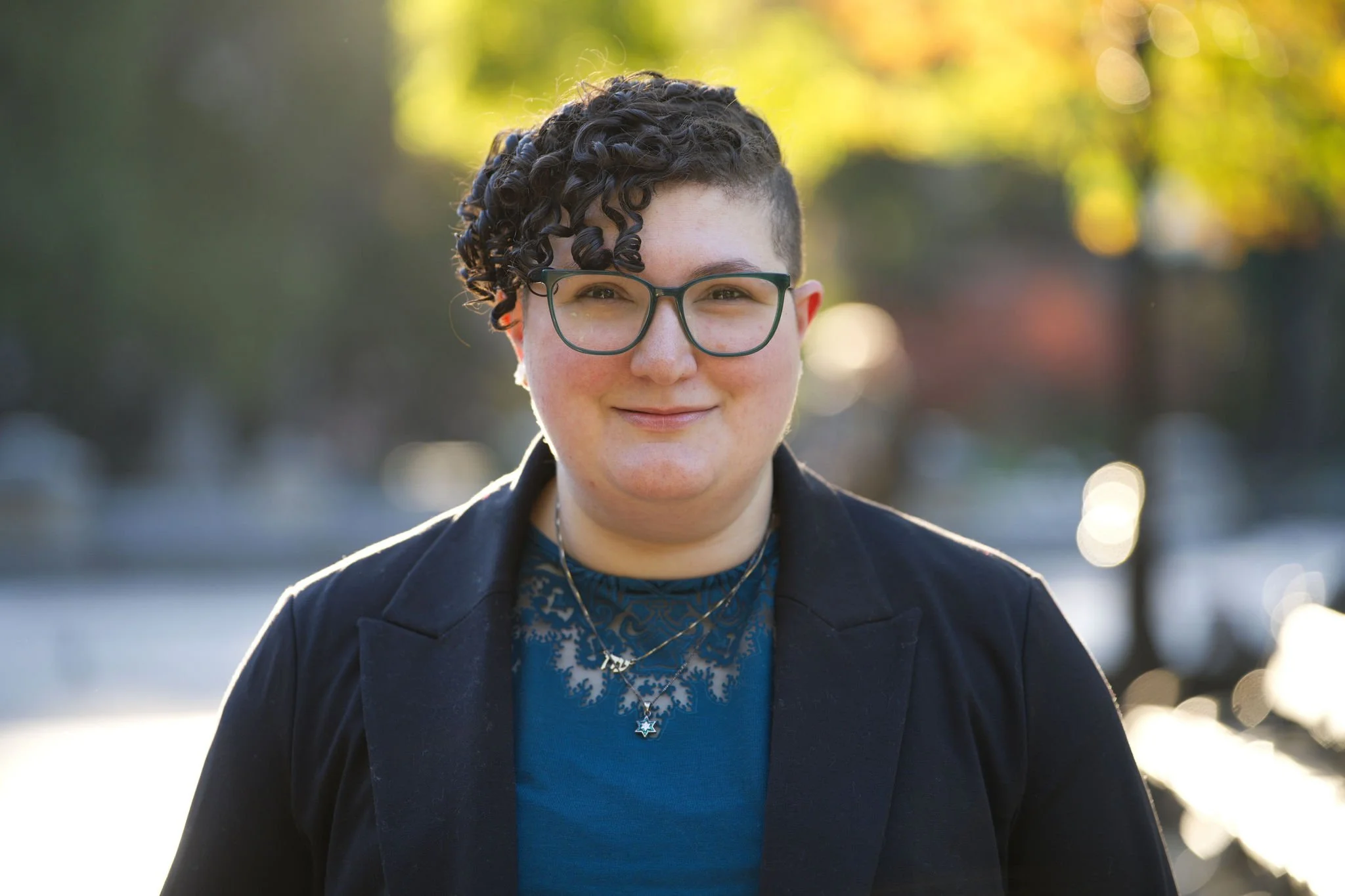 Eden has short curly hair, wearing glasses, earrings, layered necklaces, a teal top, and a black blazer. They are standing outdoors with a blurred background of trees with autumn leaves.