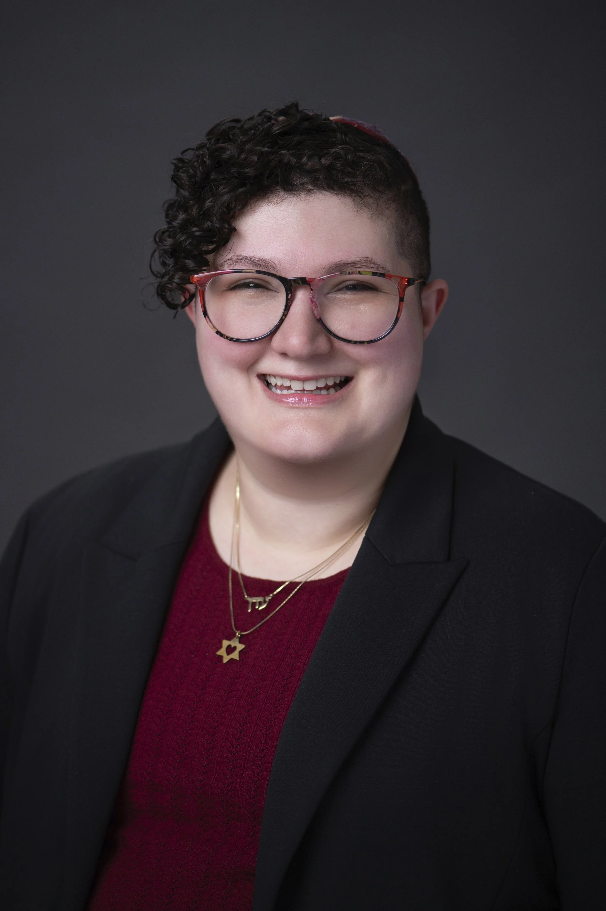 A professional headshot of Eden with short curly hair wearing glasses, a black blazer, a red sweater, and gold jewelry, smiling against a dark background.