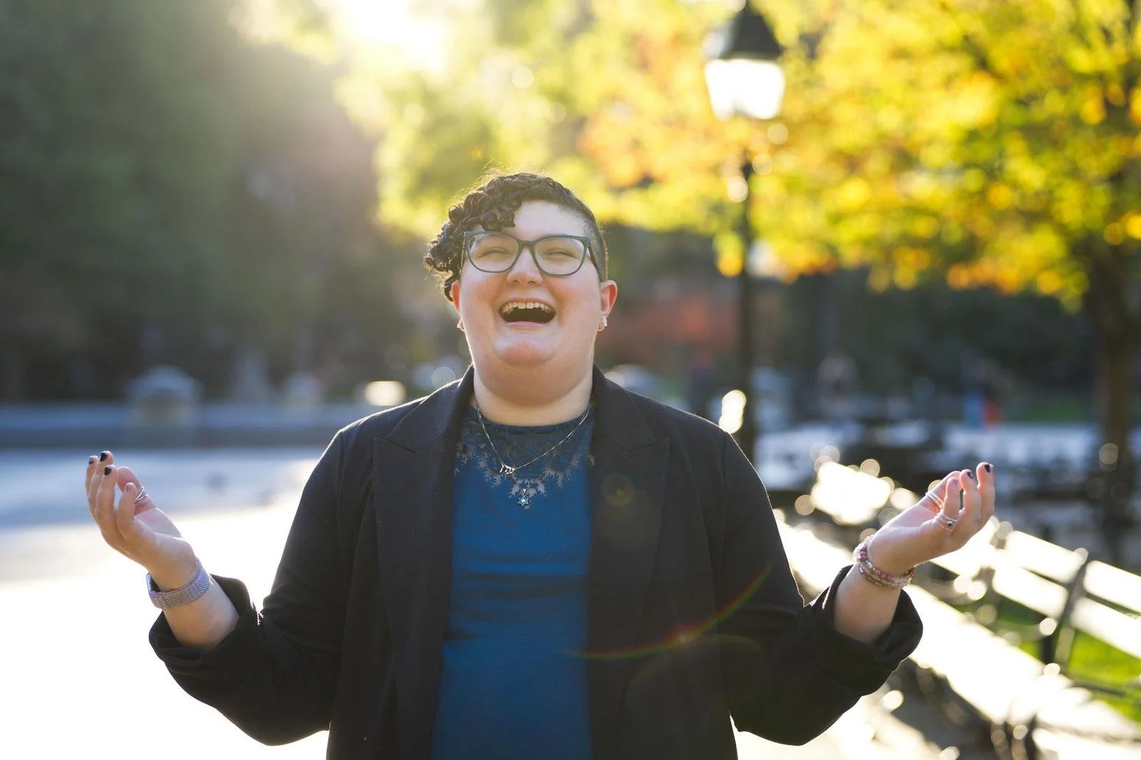Eden smiling and laughing outdoors in sunlight, wearing glasses, a black blazer, and jewelry, with arms raised in a joyful gesture, in a park with trees and benches in the background.