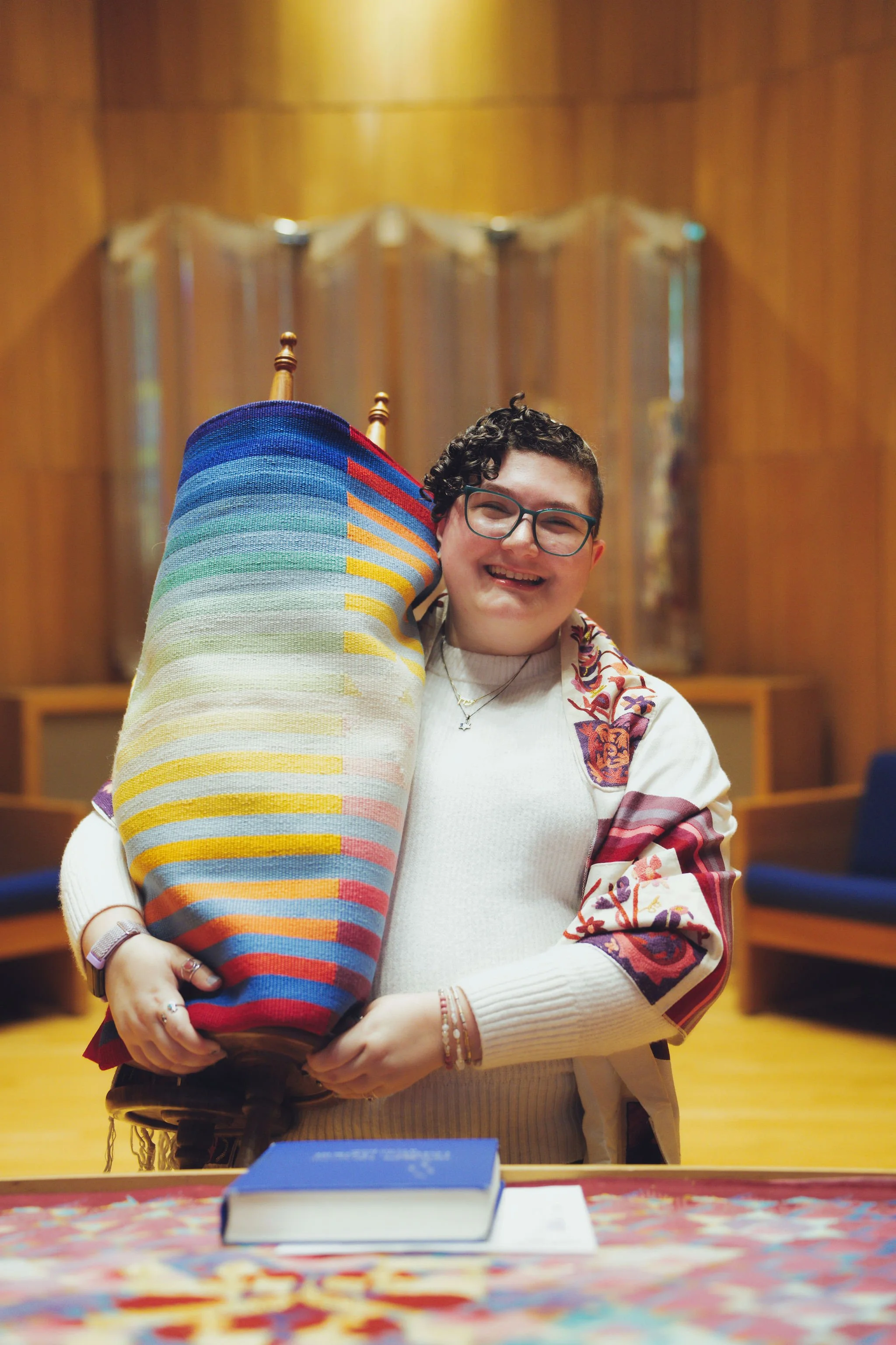 Eden smiling and holding a Torah with a colorful woven mantel in a wood-paneled room.