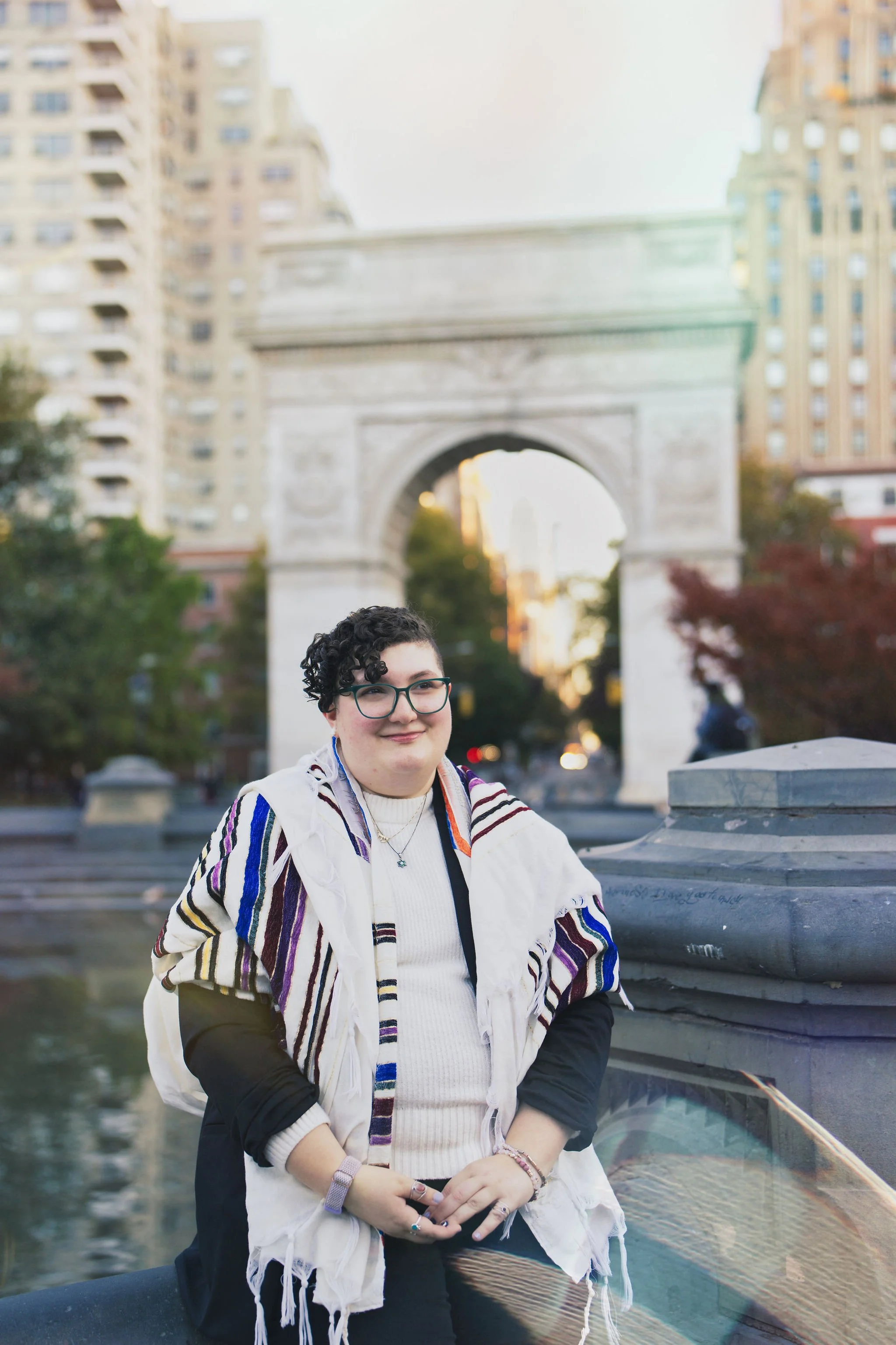 Eden is a person with glasses and short curly hair smiling, wearing a striped large prayer shawl, white sweater, and black blazer. They are sitting in front of the Washington Square Park arch, with trees and tall buildings in the background.