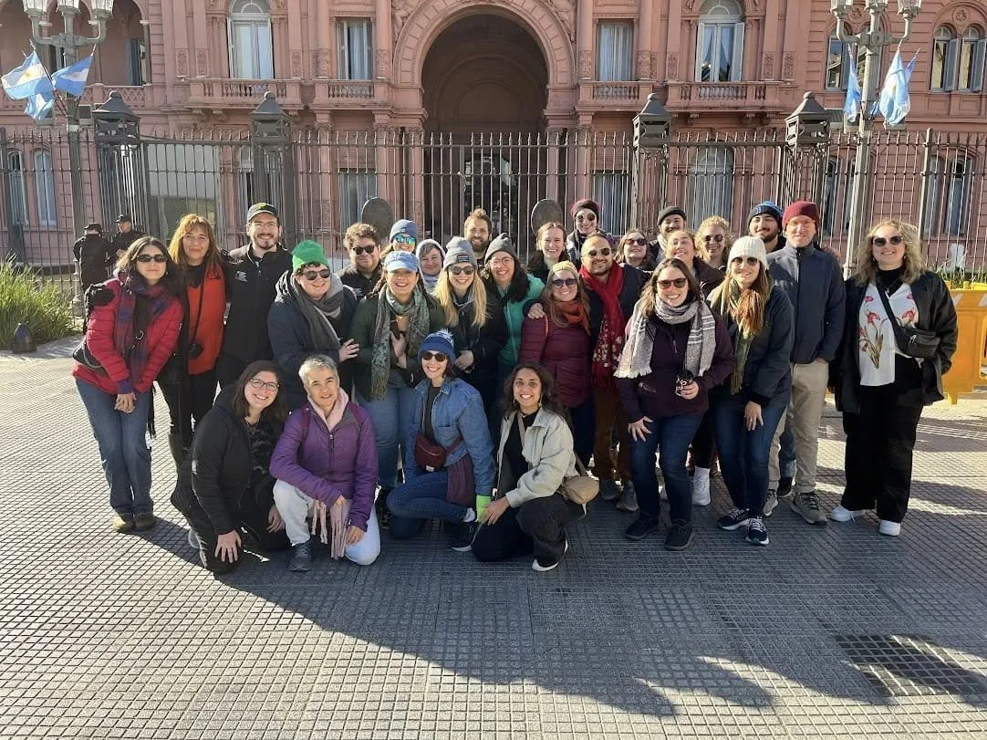Group of people posing for a photo in front of a historic pink building with iron gates.