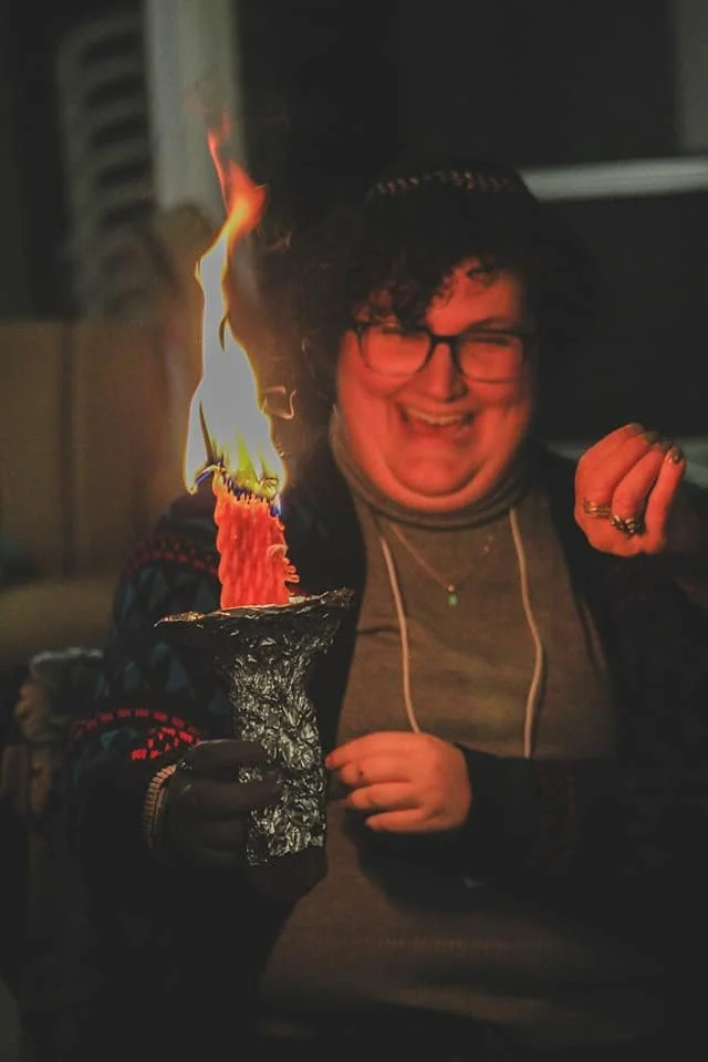 Eden smiling and holding a Havdalah candle with a large flame, in a dark indoor setting.
