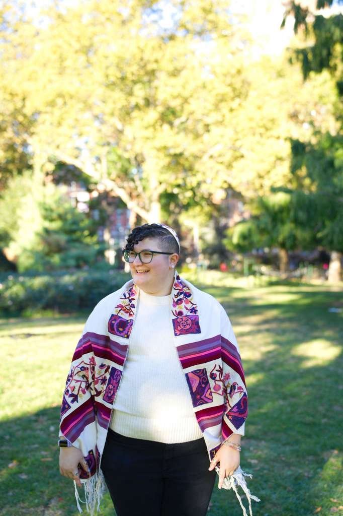 A photo of Eden who has short, curly hair and glasses, smiling in a park, wearing a white sweater and a colorful tallit, prayer shawl, with purple, pink, and red patterns, standing on grass with trees and sunlight in the background.