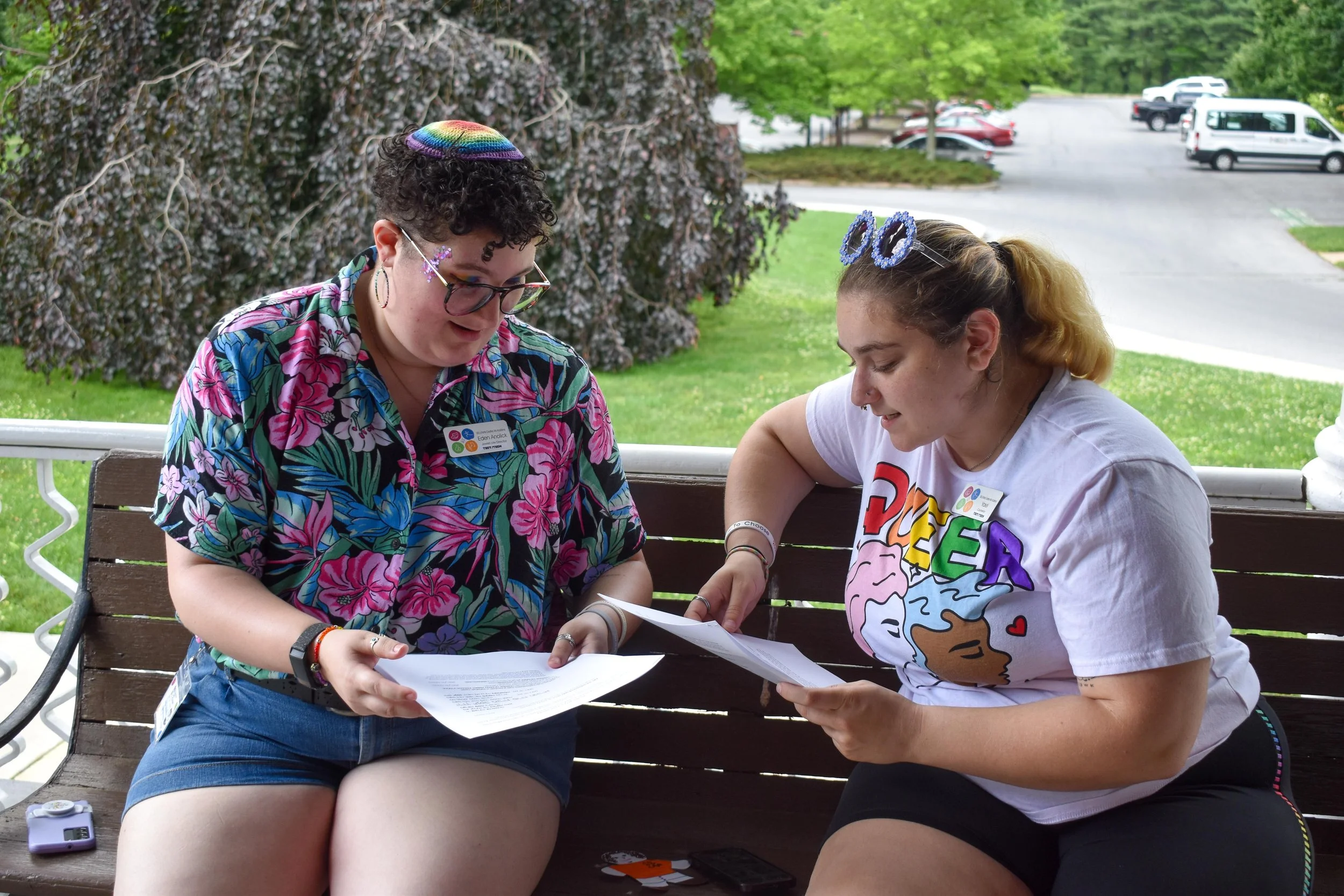 Eden and their friend, sitting on a bench, looking at papers together and smiling. One is wearing a colorful Hawaii shirt and glasses, the other a white T-shirt with colorful text and cartoon character. It is a bright, green outdoor setting.