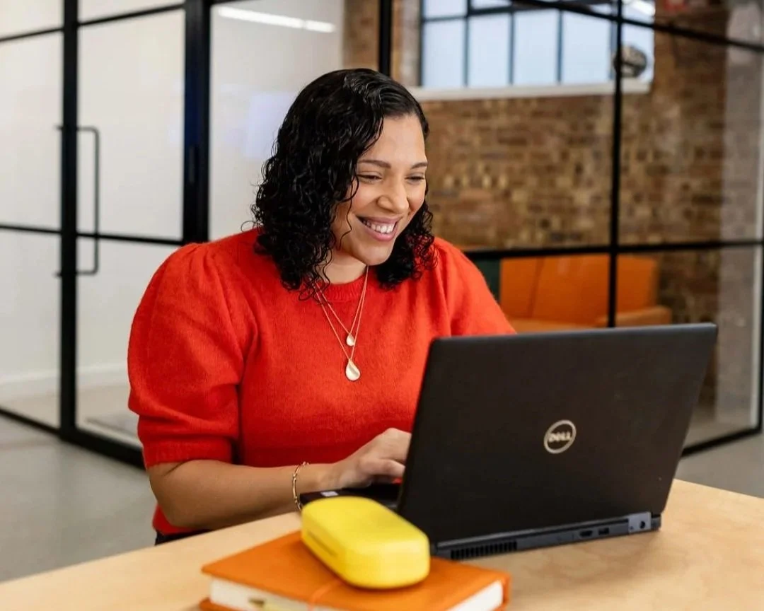 A woman with curly black hair smiling while working on a Dell laptop in an office, wearing a red sweater and jewelry, with a yellow container and an orange notebook on the desk.