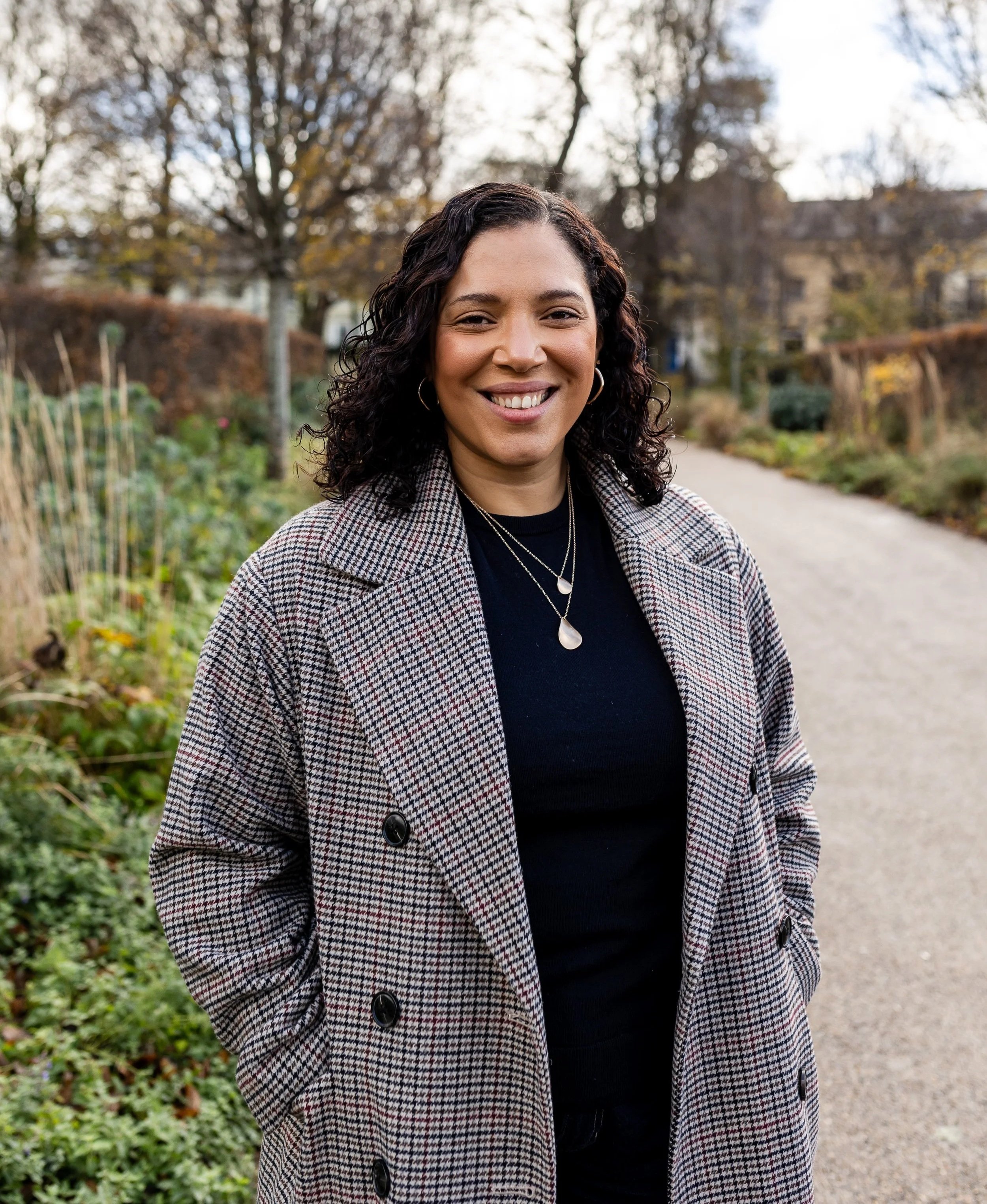 Smiling woman wearing a checkered coat and layered necklaces standing outdoors on a pathway with trees and houses in the background.