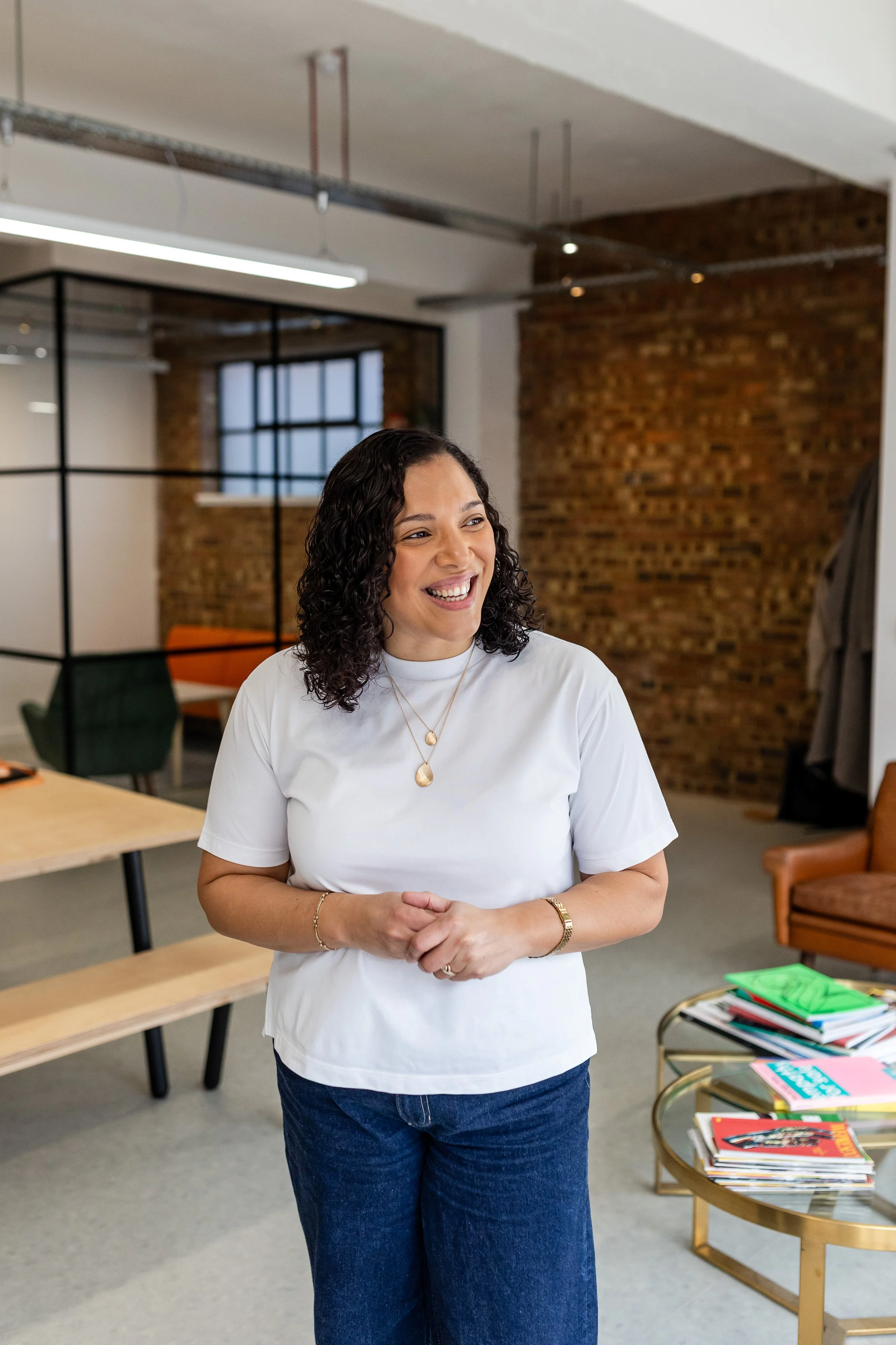 A woman with curly dark hair, wearing a white T-shirt and blue jeans, smiling in an office with exposed brick walls, a window, and a modern interior.